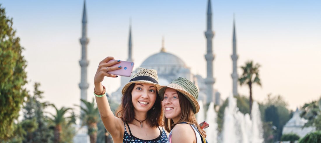 Eskapas Turkey tours Two smiling girlfriends with smart phone doing selfie in front of the Blue Mosque in Istanbul. Travel and vacation in Turkey concept