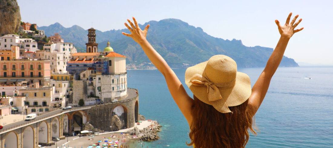 Summer holiday in Italy. Back view of young woman with straw hat and yellow dress with raised arms looking at Atrani village, Amalfi Coast, Italy